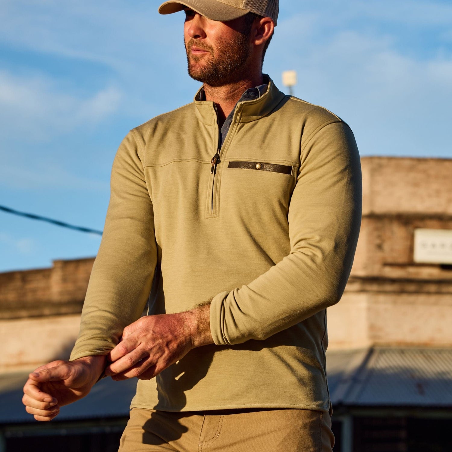 A person wearing a beige cap and Tom Beckbes Tucker Fleece Quarter Zip adjusts their sleeve outdoors against a blue sky.