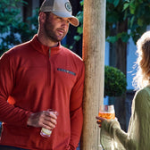 A man in a red Tucker Fleece Quarter Zip by Tom Beckbe chats with a blonde by a wooden post, both enjoying drinks.