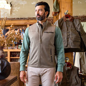 A man wearing a gray vest and green checkered shirt stands in a rustic clothing store with jackets hanging behind him.