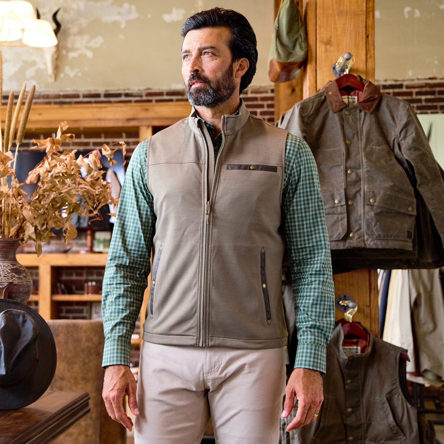 A man wearing a gray vest and green checkered shirt stands in a rustic clothing store with jackets hanging behind him.