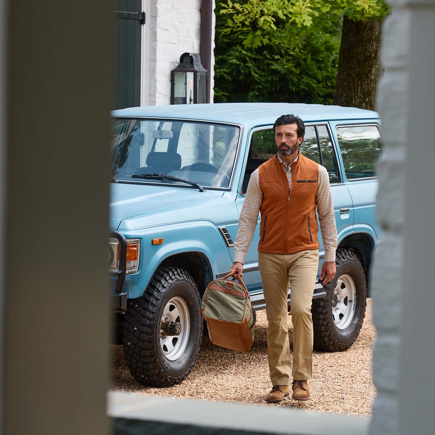 Man in outdoor clothing carrying a duffel bag walking past a blue SUV parked on a gravel driveway.