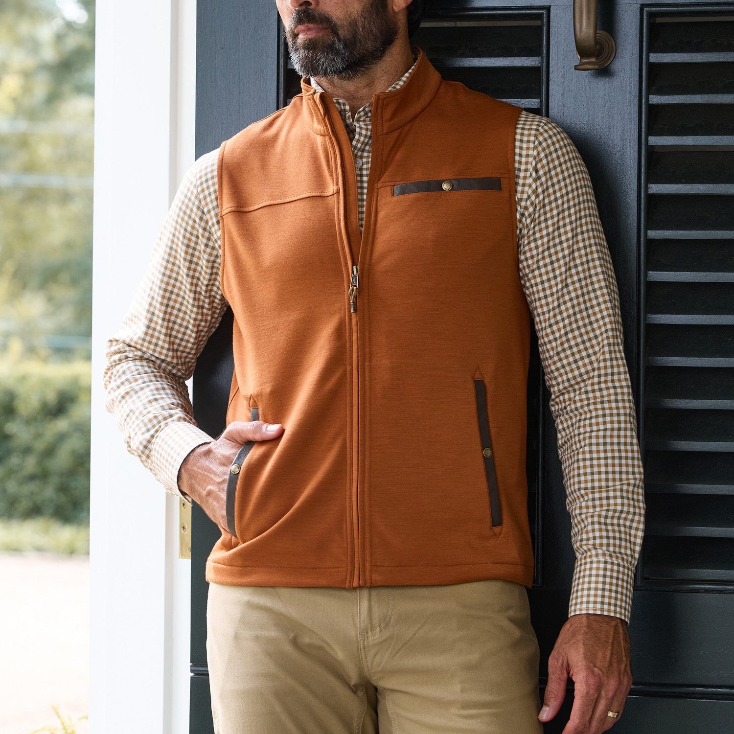 Man wearing an orange zip-up vest over a checked shirt and khaki pants, standing in front of a dark door.