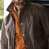 Man wearing a brown waxed jacket over an orange zip-up sweater and a checkered collared shirt.