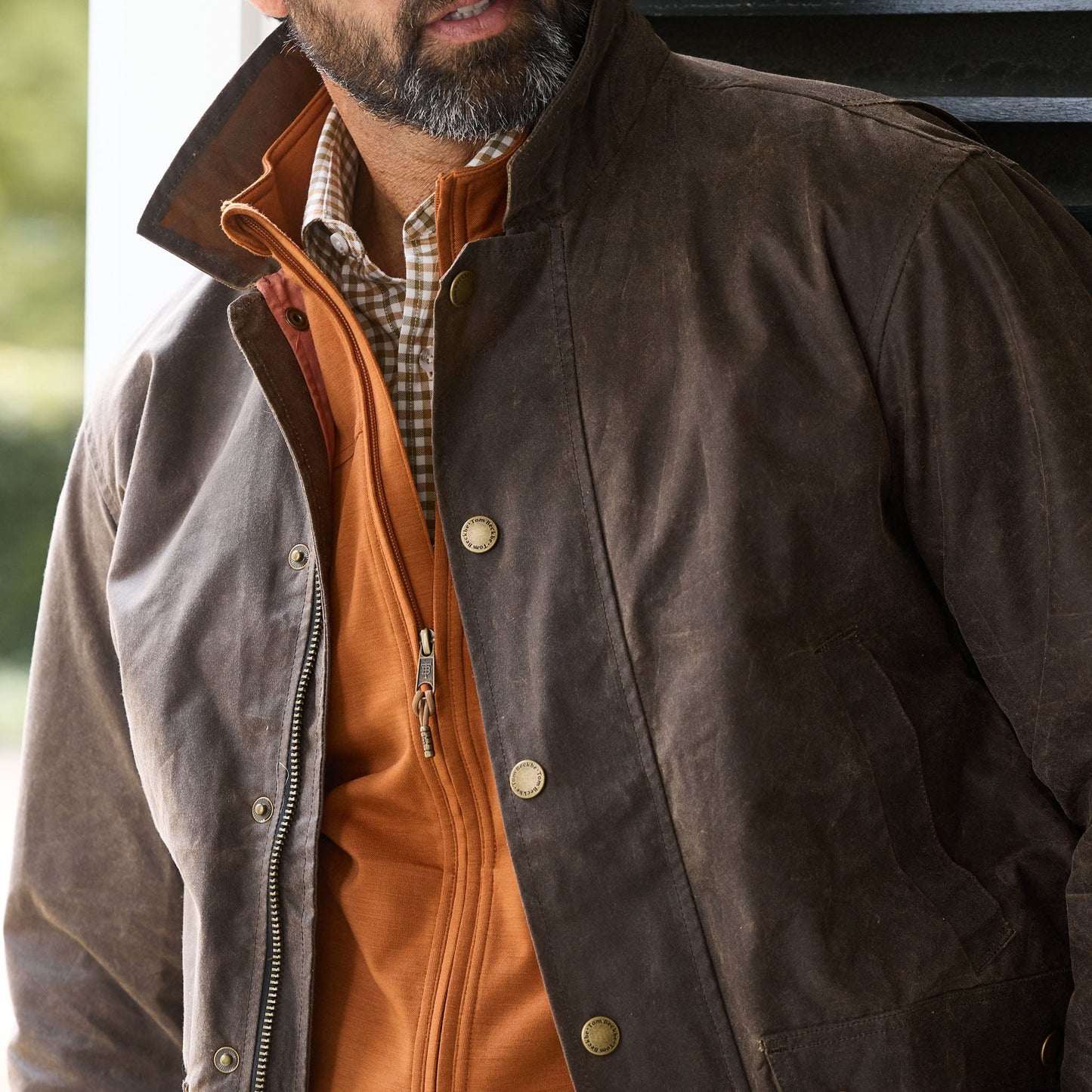 Man wearing a brown waxed jacket over an orange zip-up sweater and a checkered collared shirt.
