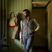 Man in a gray vest and pants stands by a door, looking out, with a coat and bag hanging on the wall behind him.