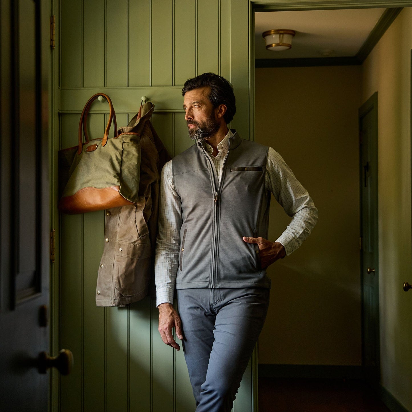 Man in a gray vest and pants stands by a door, looking out, with a coat and bag hanging on the wall behind him.