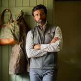 Man with beard wearing a gray vest and striped shirt stands with arms crossed in front of a coat rack with bags and a jacket.
