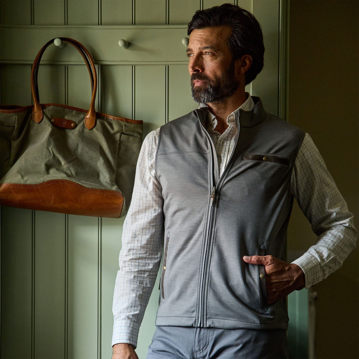 Man wearing a gray zip-up vest and plaid shirt stands indoors next to a wall with a hanging green and brown bag.
