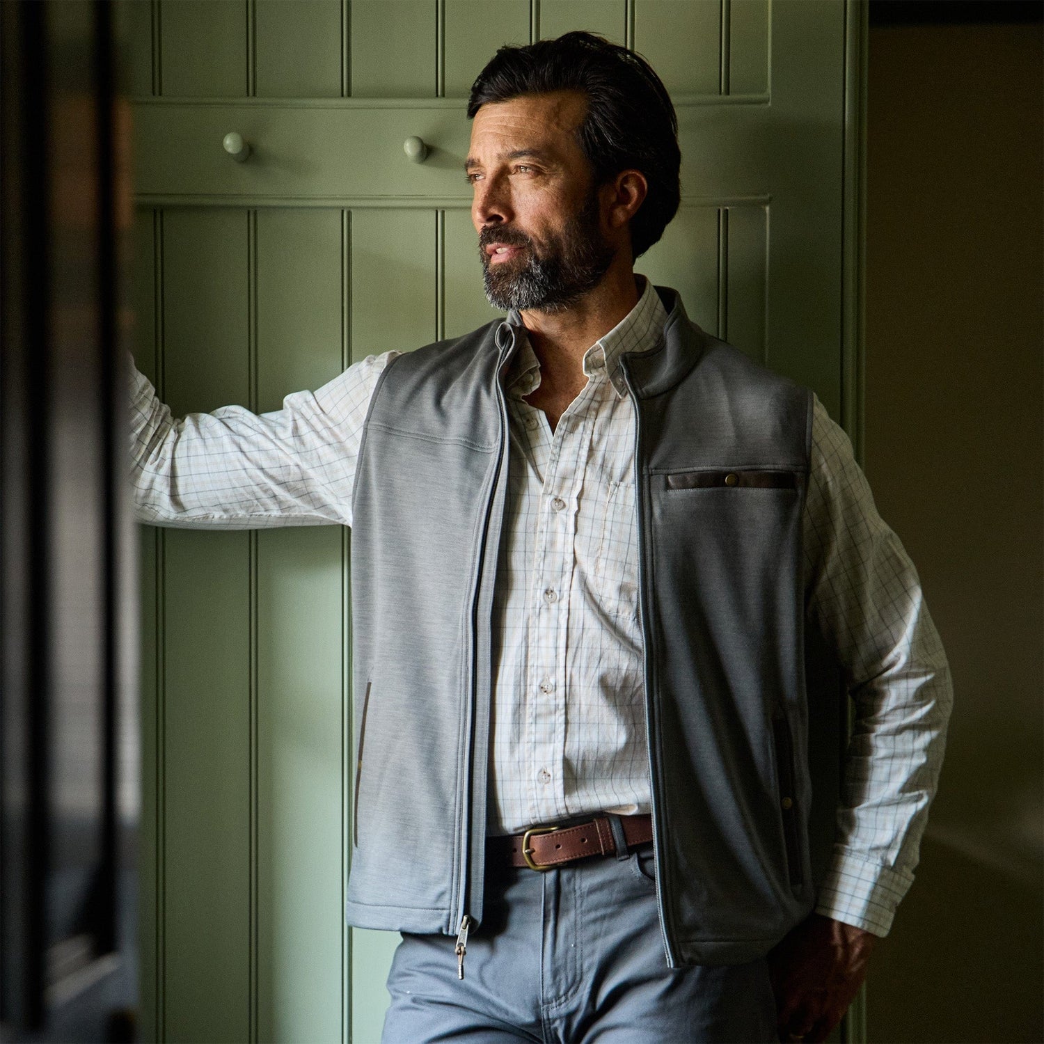 Man with a beard wearing a gray vest and striped shirt stands indoors, looking out a window with his hand on the frame.