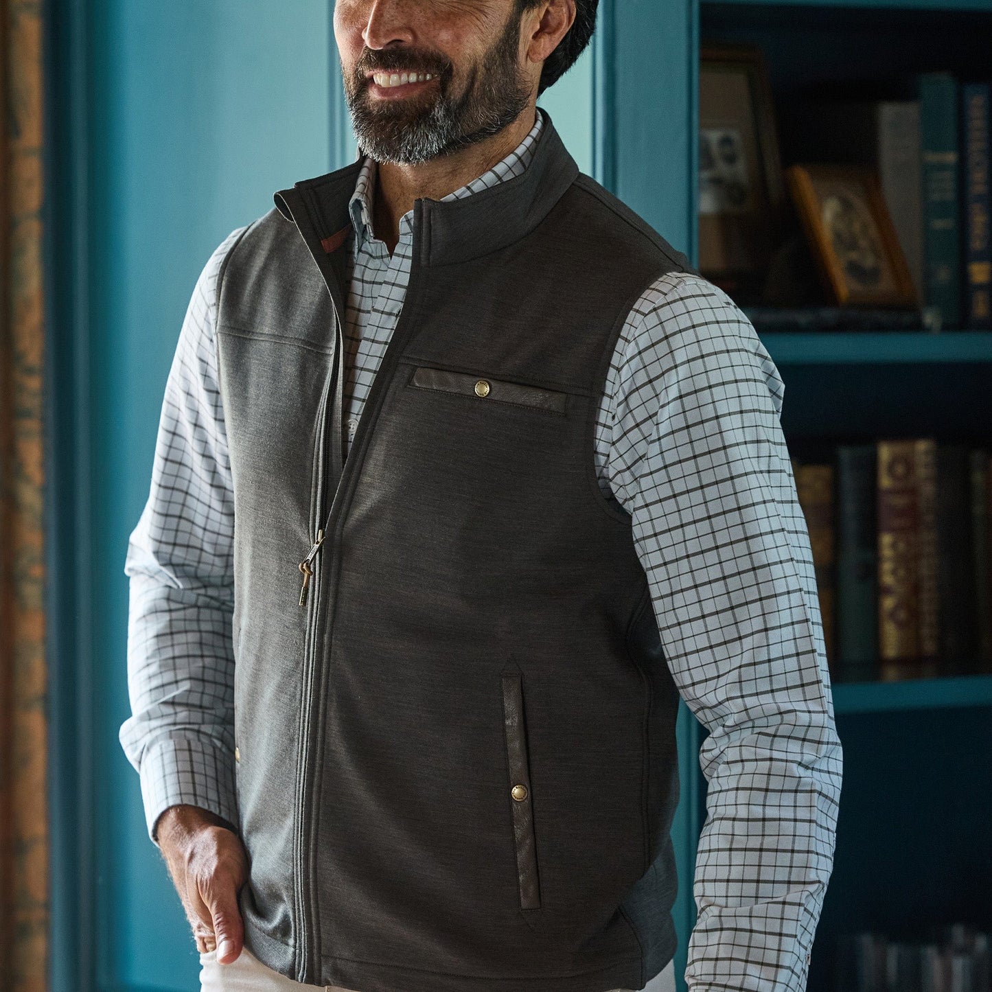 Man wearing a gray zip-up vest over a white checkered shirt, standing indoors near shelves with books and decor.