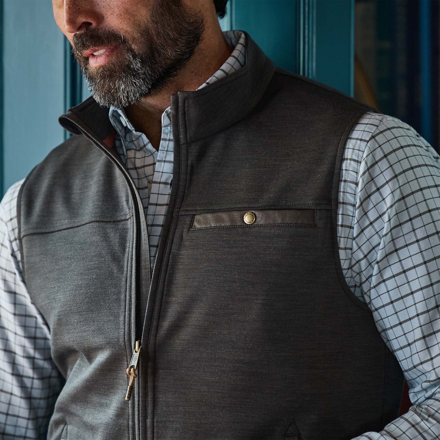A man wearing a gray zip-up vest over a white plaid shirt stands in front of a blue door.