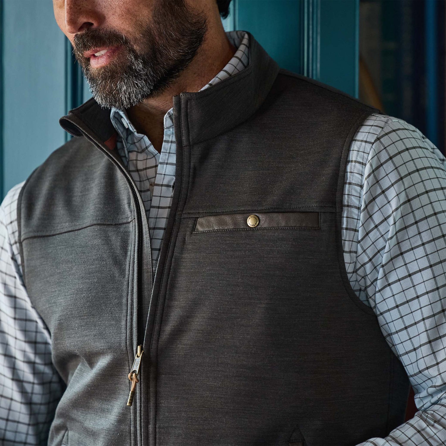A man wearing a gray zip-up vest over a white plaid shirt stands in front of a blue door.