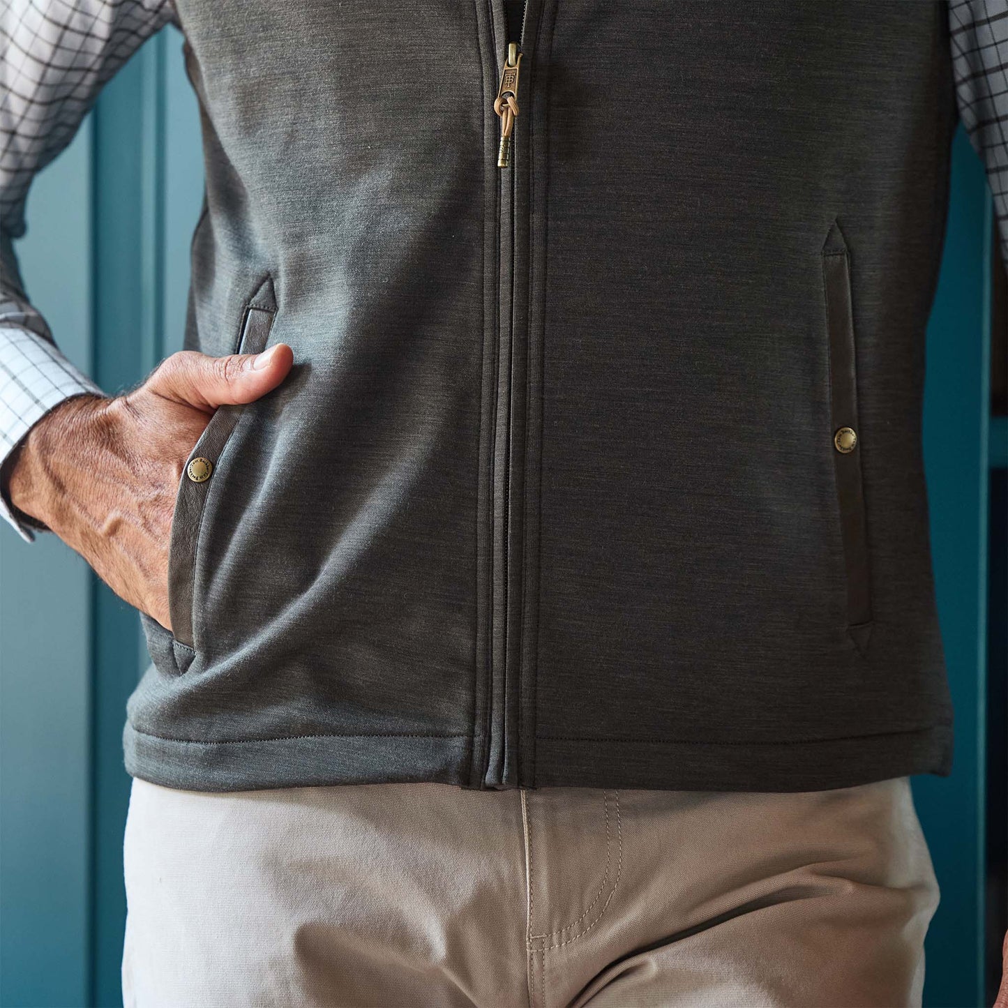 Person wearing a zip-up vest over a checkered shirt and beige pants, with one hand in the vest pocket.