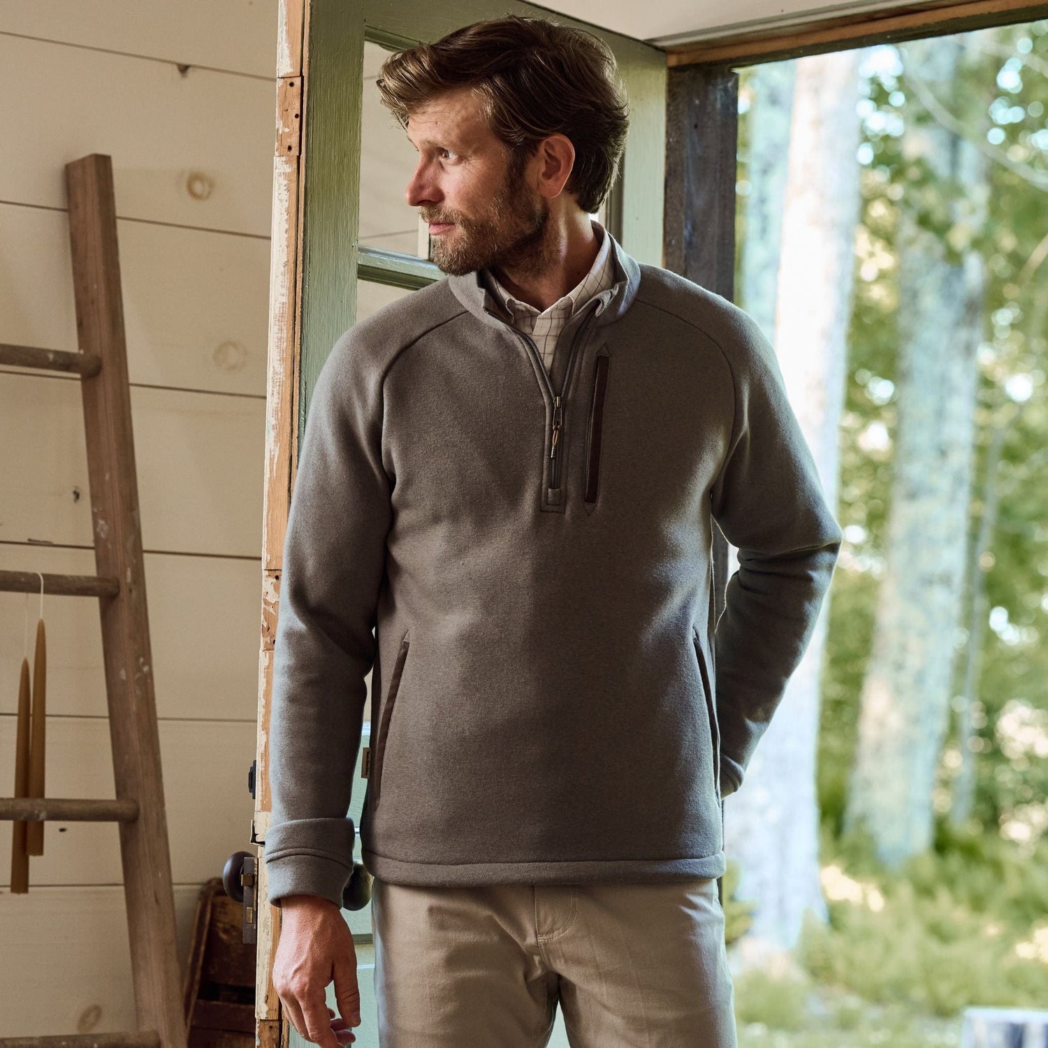 A man in a Tom Beckbe Warren Fleece Quarter Zip stands in a doorway, with a ladder leaning against the nearby wooden wall.