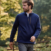 A man wearing a Tom Beckbe Warren Fleece Quarter Zip strolls outdoors on a sunny day with trees in the background.