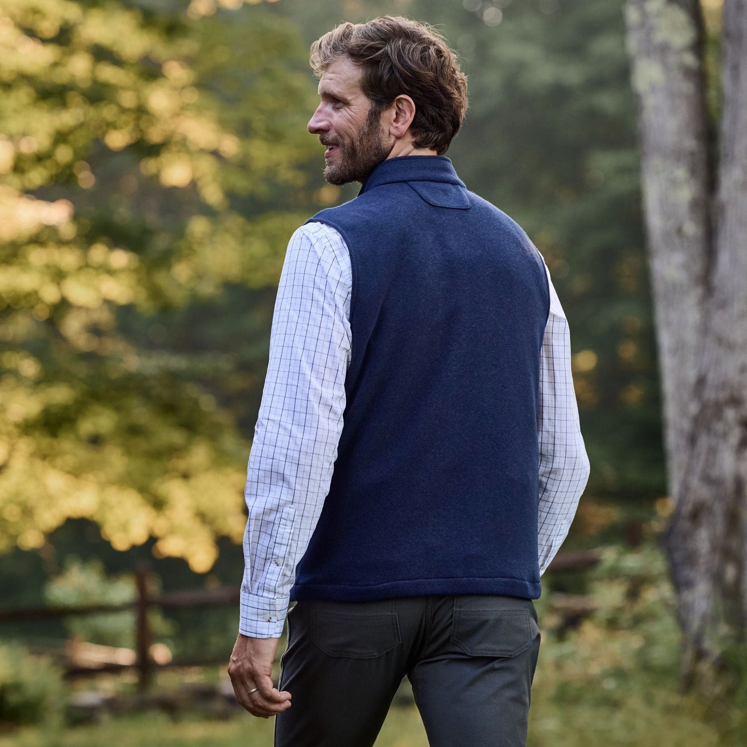 Man in a Tom Beckbe Warren Fleece Vest over a plaid shirt, walking outdoors with trees and a fence behind him.
