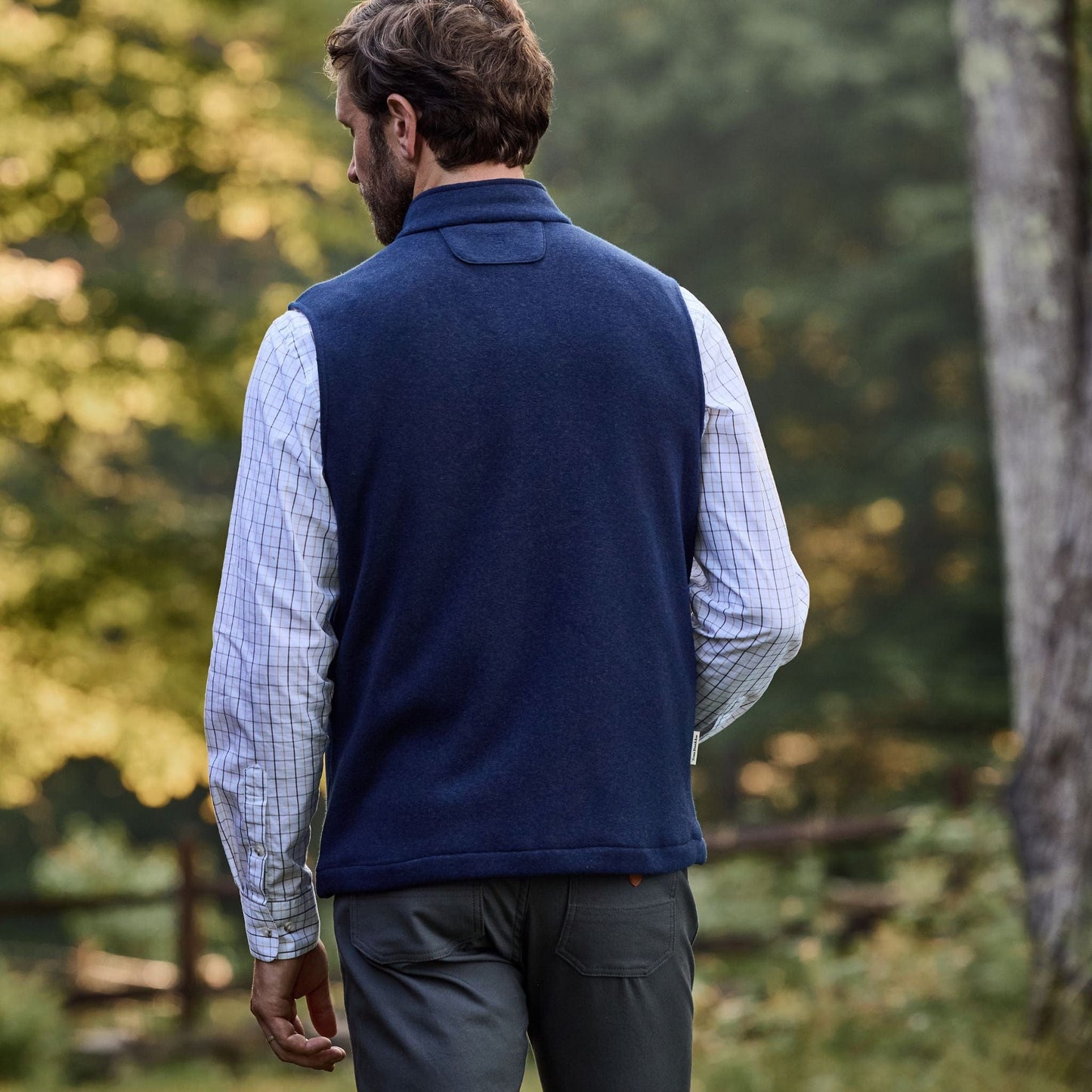 Man wearing a Tom Beckbe Warren Fleece Vest and plaid shirt outdoors, facing away with blurred green trees behind.
