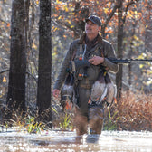 A man in a Tom Beckbe DU Waterfowl Strap Vest stands in shallow water with a rifle and ducks, surrounded by autumn trees.