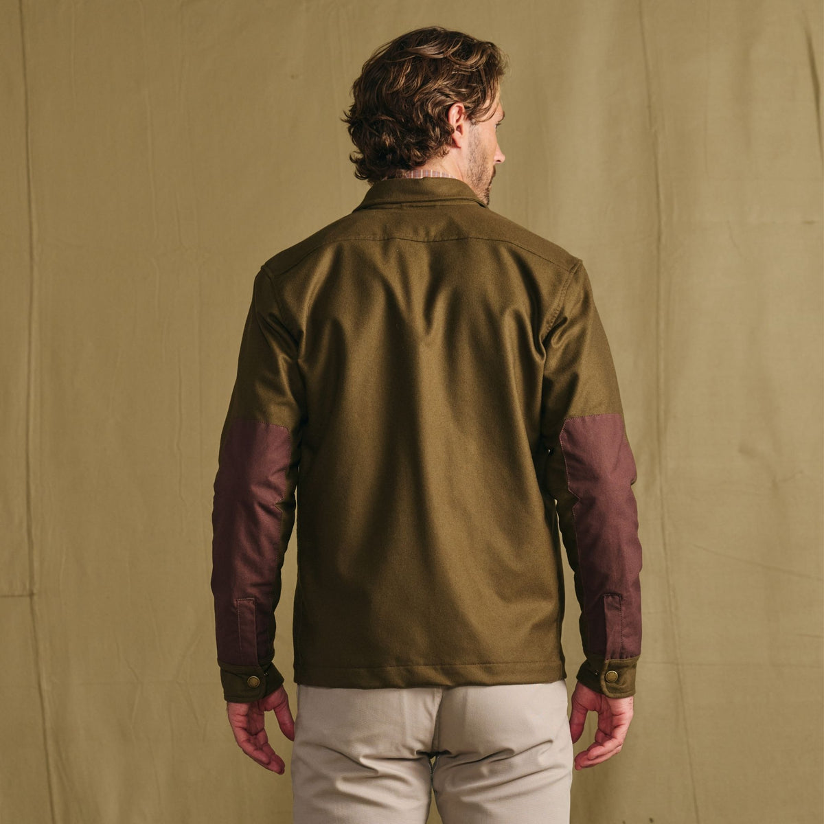 A man, back to camera, wears the Tom Beckbe Westport Wool Overshirt in olive green with brown elbow patches and light pants.