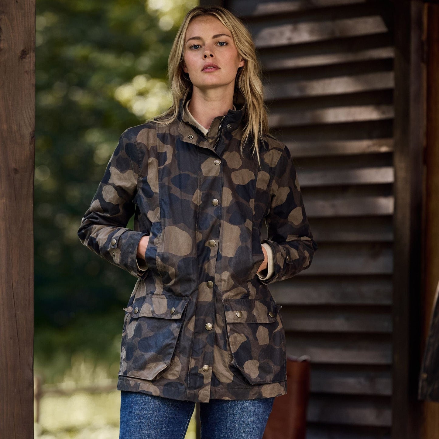 A woman stands outdoors in a Tom Beckbe Womens Blakeley Jacket with a camo pattern, paired with jeans. She leans against a wooden structure, surrounded by blurred greenery, with her hands in her pockets.