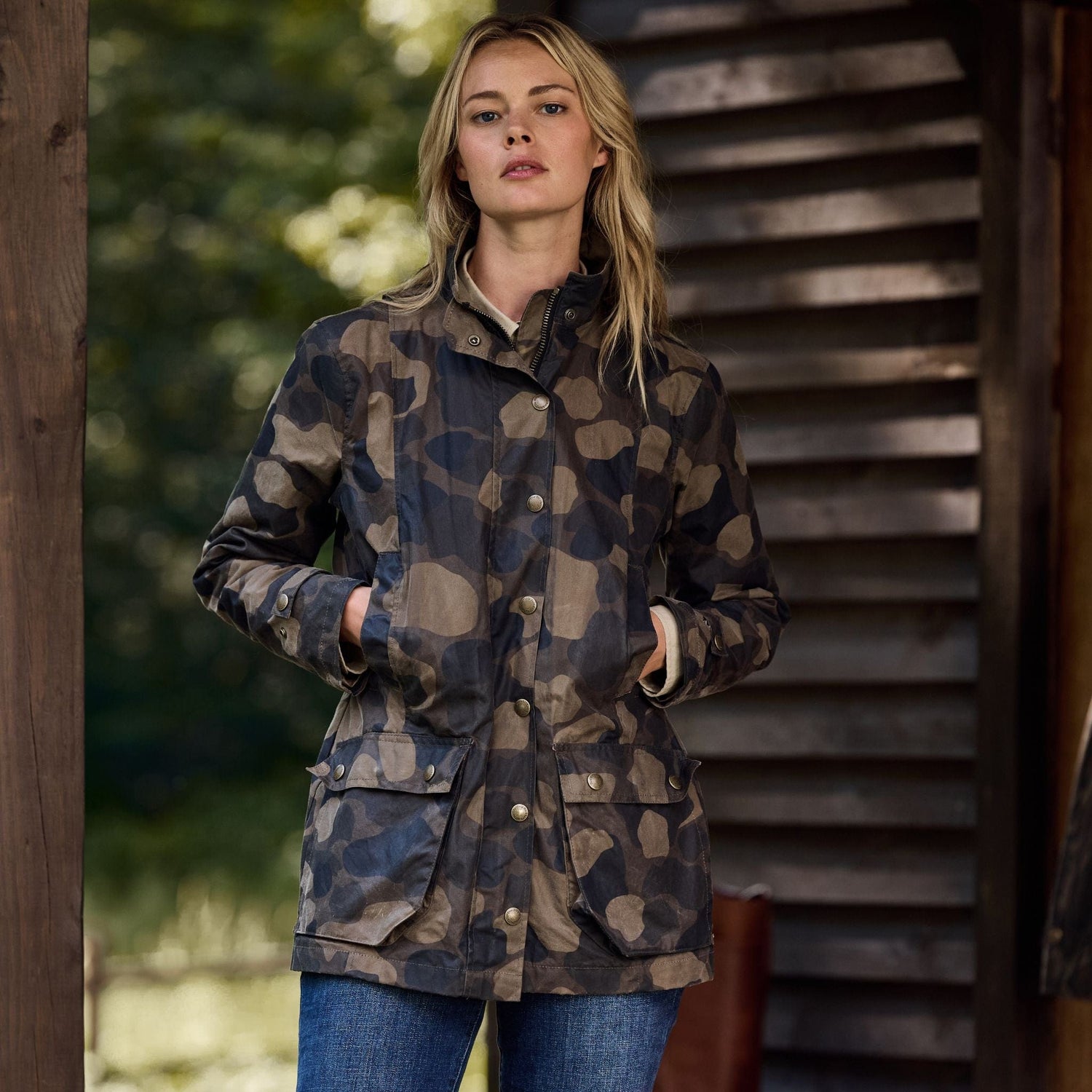 A woman stands outdoors in a Tom Beckbe Womens Blakeley Jacket with a camo pattern, paired with jeans. She leans against a wooden structure, surrounded by blurred greenery, with her hands in her pockets.