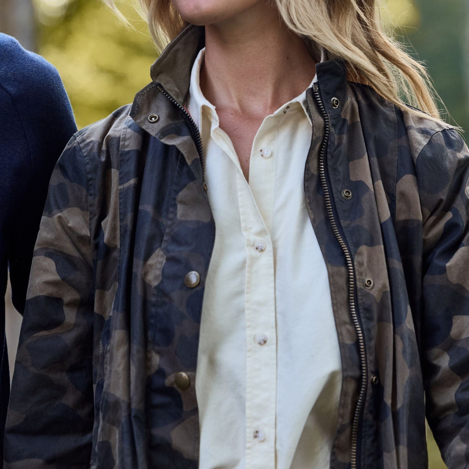 Wearing a Womens Blakeley Jacket by Tom Beckbe over a white shirt, a person strolls through blurred greenery with loose hair, capturing a casual and relaxed moment.