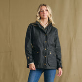 Woman wears the Tom Beckbe Women’s Blakeley Jacket with blue jeans against a beige background.