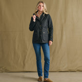 Woman indoors in Tom Beckbe Women’s Blakeley Jacket, blue jeans, and brown shoes with a tan fabric backdrop.