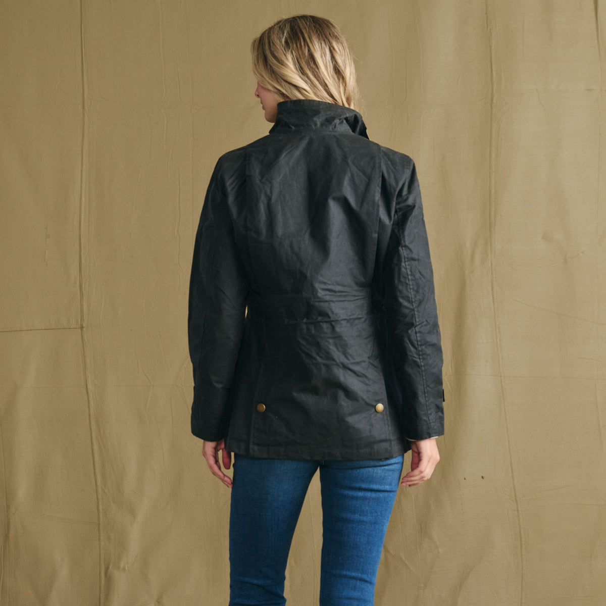 A person wearing the Women’s Blakeley Jacket by Tom Beckbe and blue jeans faces a tan fabric backdrop, shown from behind.
