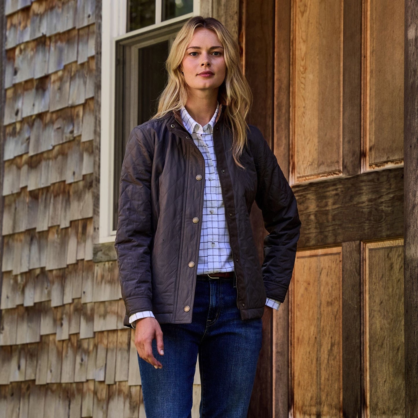 A person in a Tom Beckbe Womens Camden Quilted Jacket stands by a wooden building with a window and closed door.