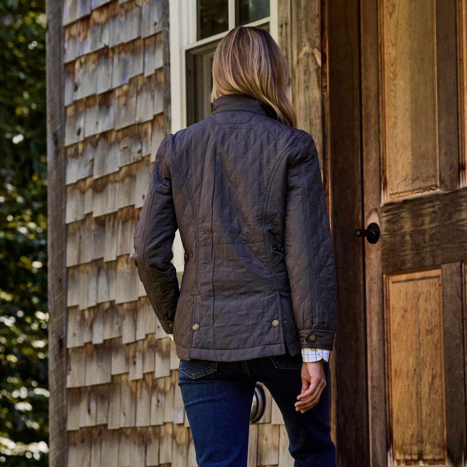 A person in a Tom Beckbe Womens Camden Quilted Jacket stands at a wooden door on a shingled building.