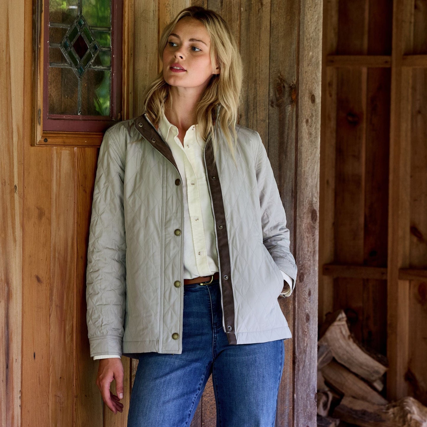 Woman in a Tom Beckbe Camden Jacket with premium insulation, leaning against a wooden wall with firewood and window.