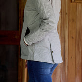 Woman in Tom Beckbes light gray Womens Camden Quilted Jacket with hands in pockets against a wooden wall.