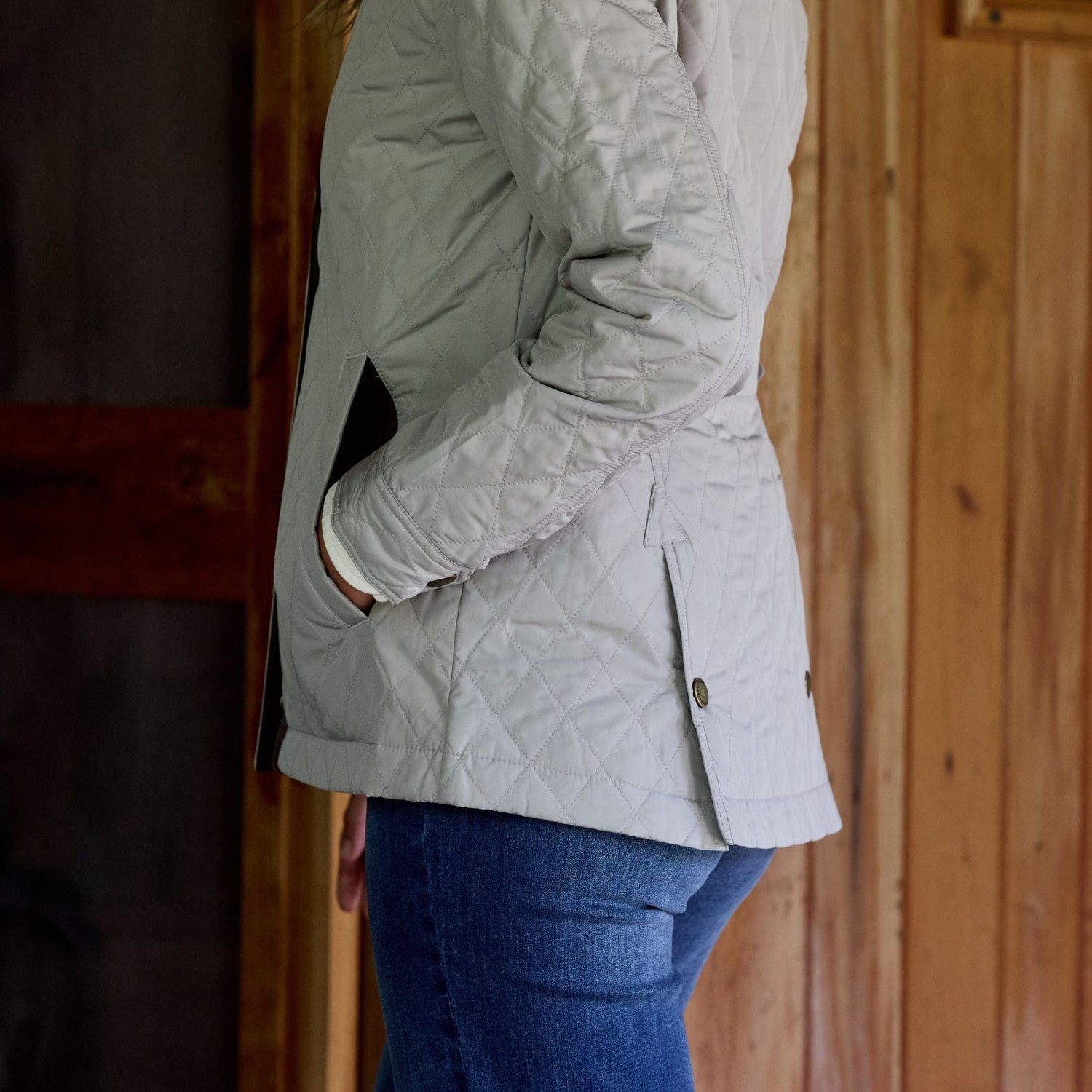 Woman in Tom Beckbes light gray Womens Camden Quilted Jacket with hands in pockets against a wooden wall.