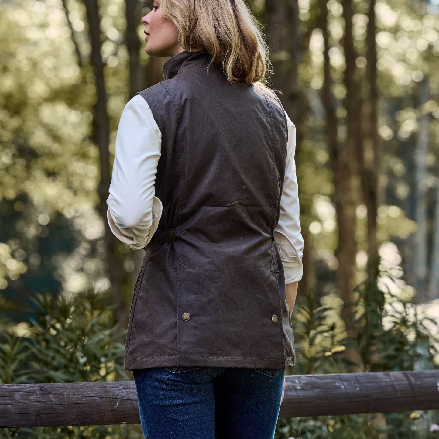 A woman in a Tom Beckbe Womens Fairmont Vest and white shirt stands by a wooden fence, facing a forest.