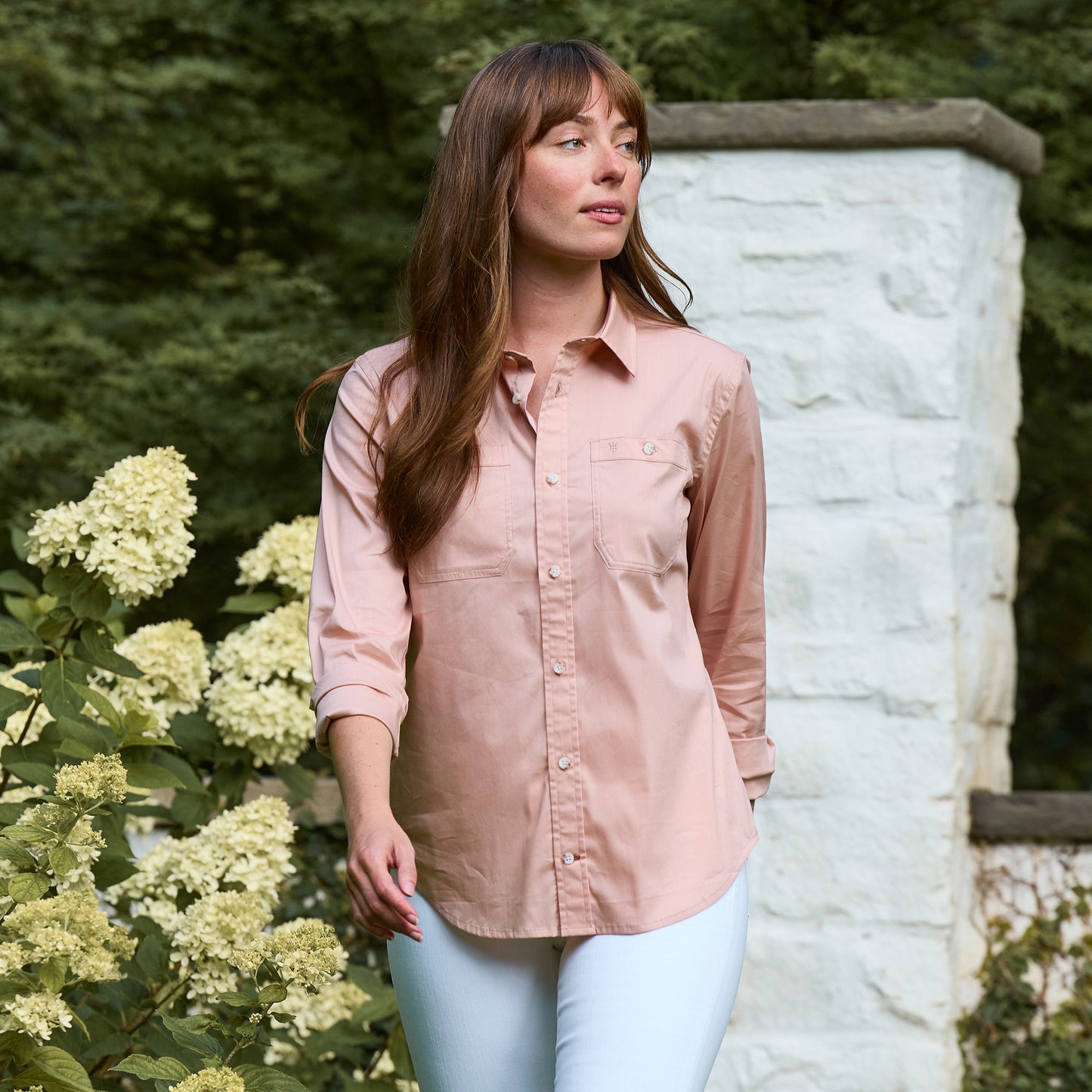A woman in the Tom Beckbe Women's Tidewater Shirt stands outdoors near white flowers and a stone wall.