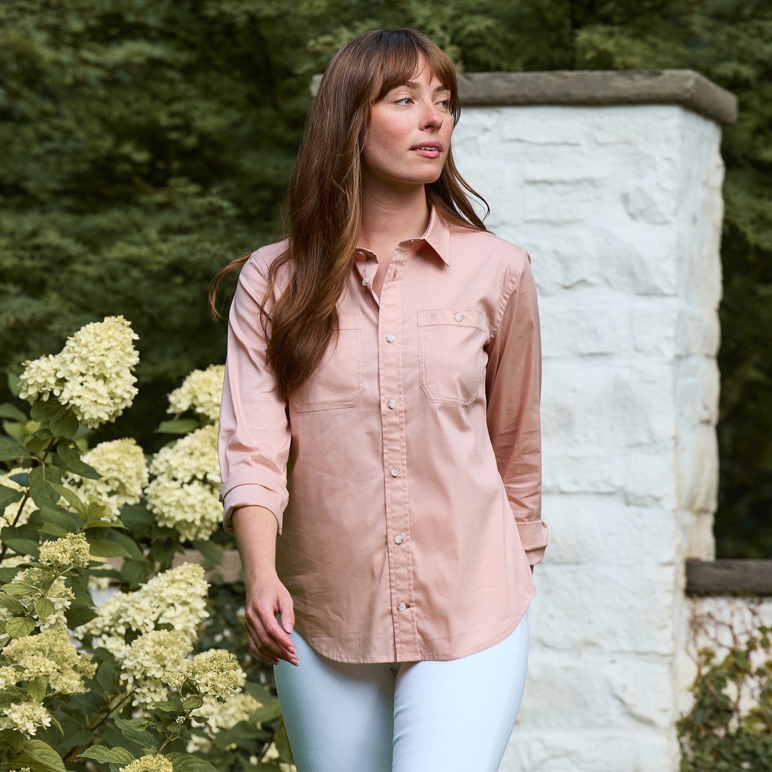 A woman in the Tom Beckbe Women's Tidewater Shirt stands outdoors near white flowers and a stone wall.