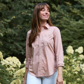 A woman in a Tom Beckbe Women's Tidewater Shirt and white pants stands outdoors among flowering bushes and greenery.