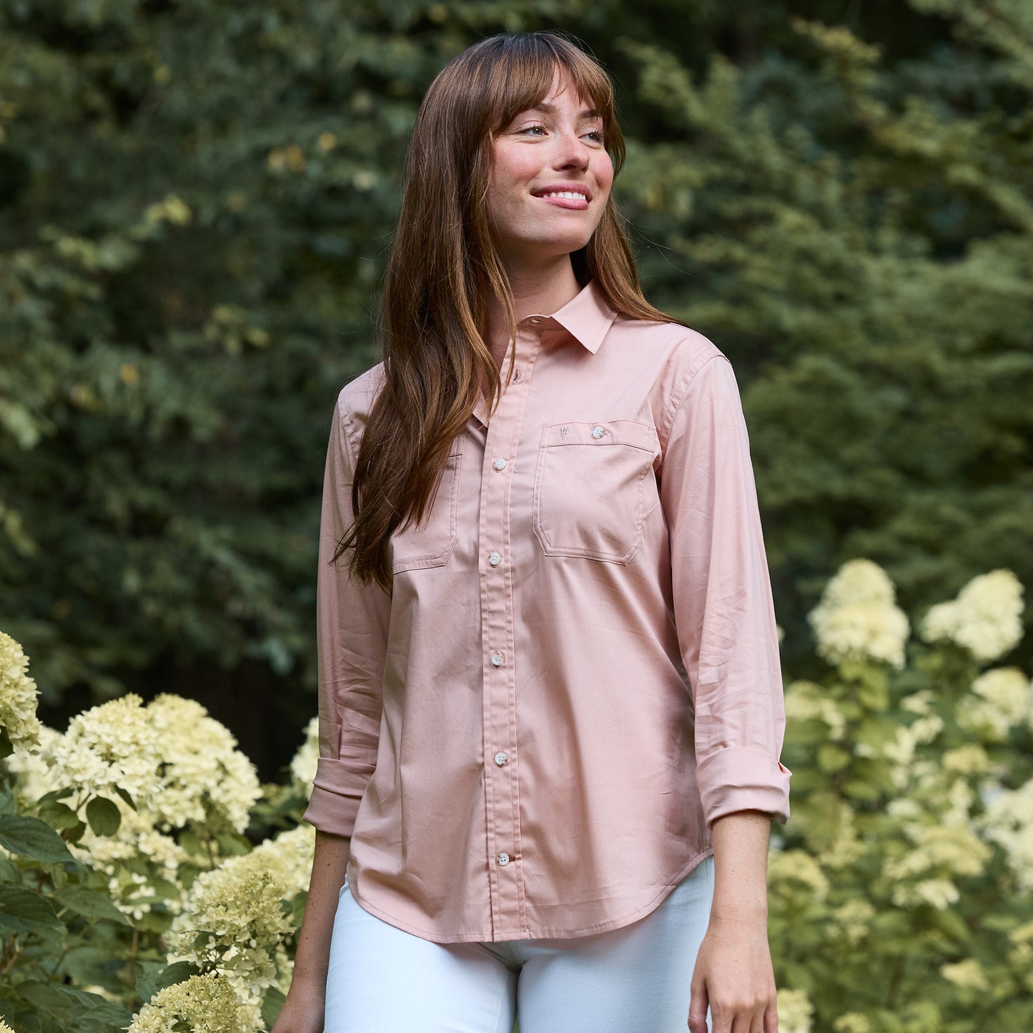 A woman in a Tom Beckbe Women's Tidewater Shirt and white pants stands outdoors among flowering bushes and greenery.