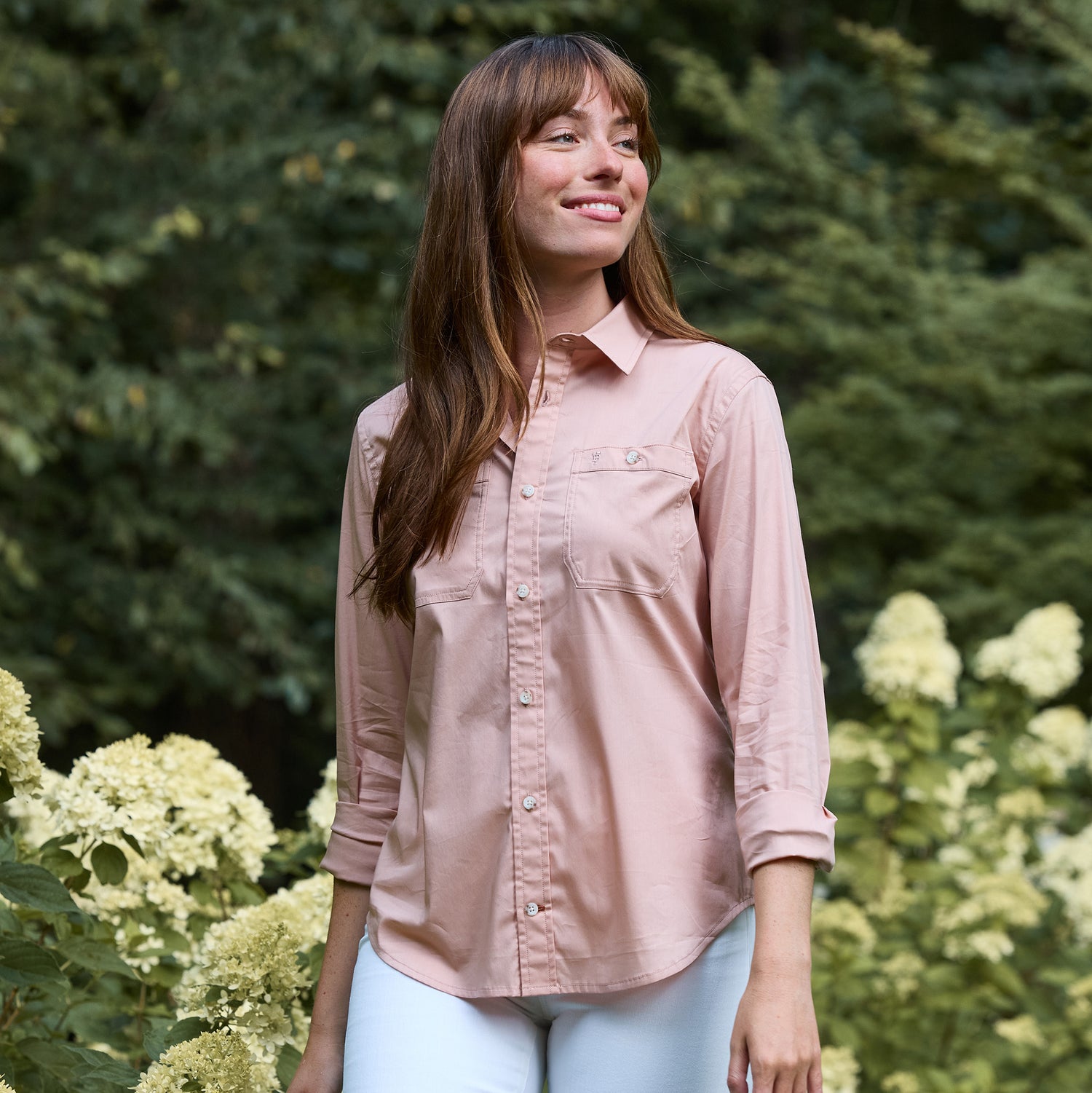 A woman in a Tom Beckbe Women's Tidewater Shirt and white pants stands outdoors among flowering bushes and greenery.