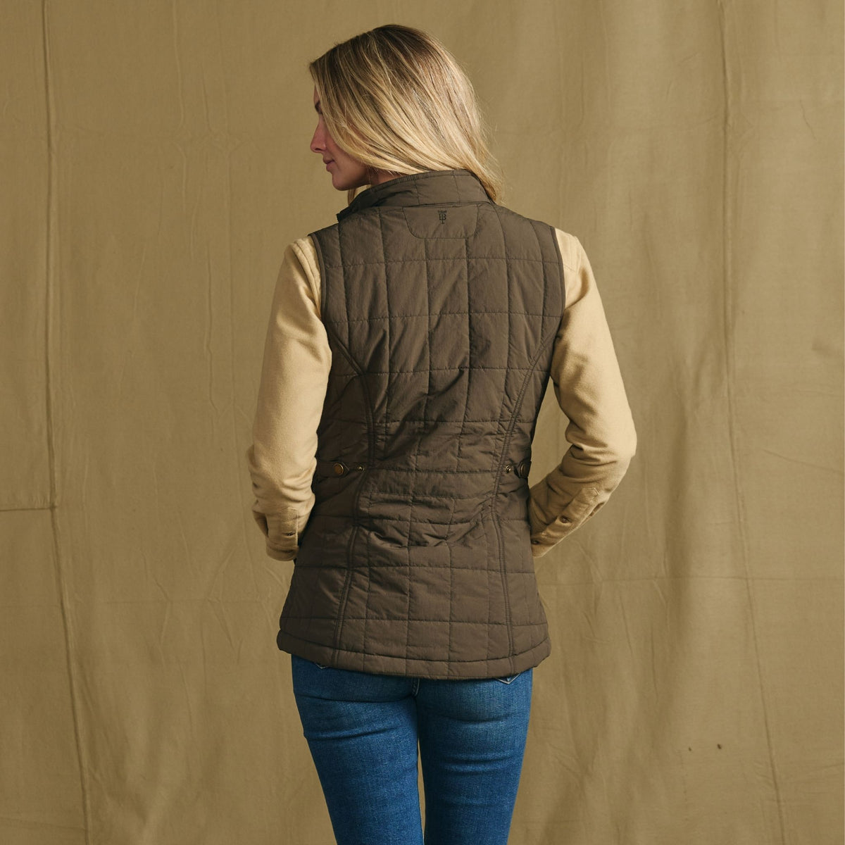 Person with back to camera wears Tom Beckbe Women’s Tyler Quilted Vest, tan shirt, and blue jeans.