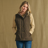 Woman wears Tom Beckbe Women’s Tyler Quilted Vest in brown over a tan shirt and jeans, styled against a beige backdrop.