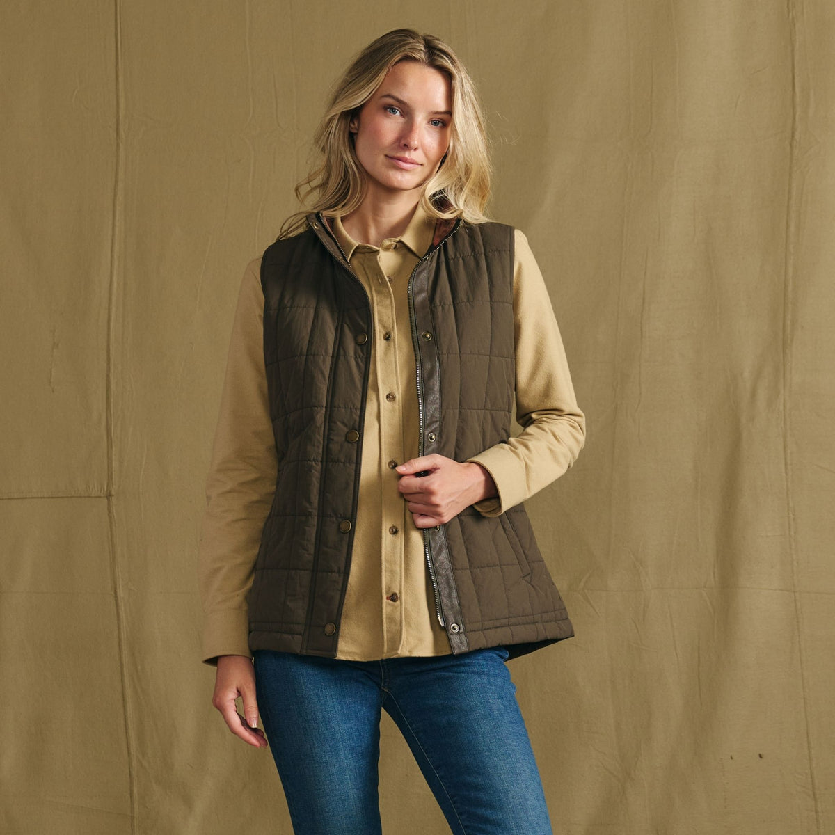 Woman wears the Tom Beckbe Women’s Tyler Quilted Vest over a tan shirt and jeans, standing against a tan background.