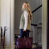 Woman stands indoors with stairs behind, wearing Tom Beckbe Women’s Tyler Quilted Vest, jeans, and holding a leather bag.