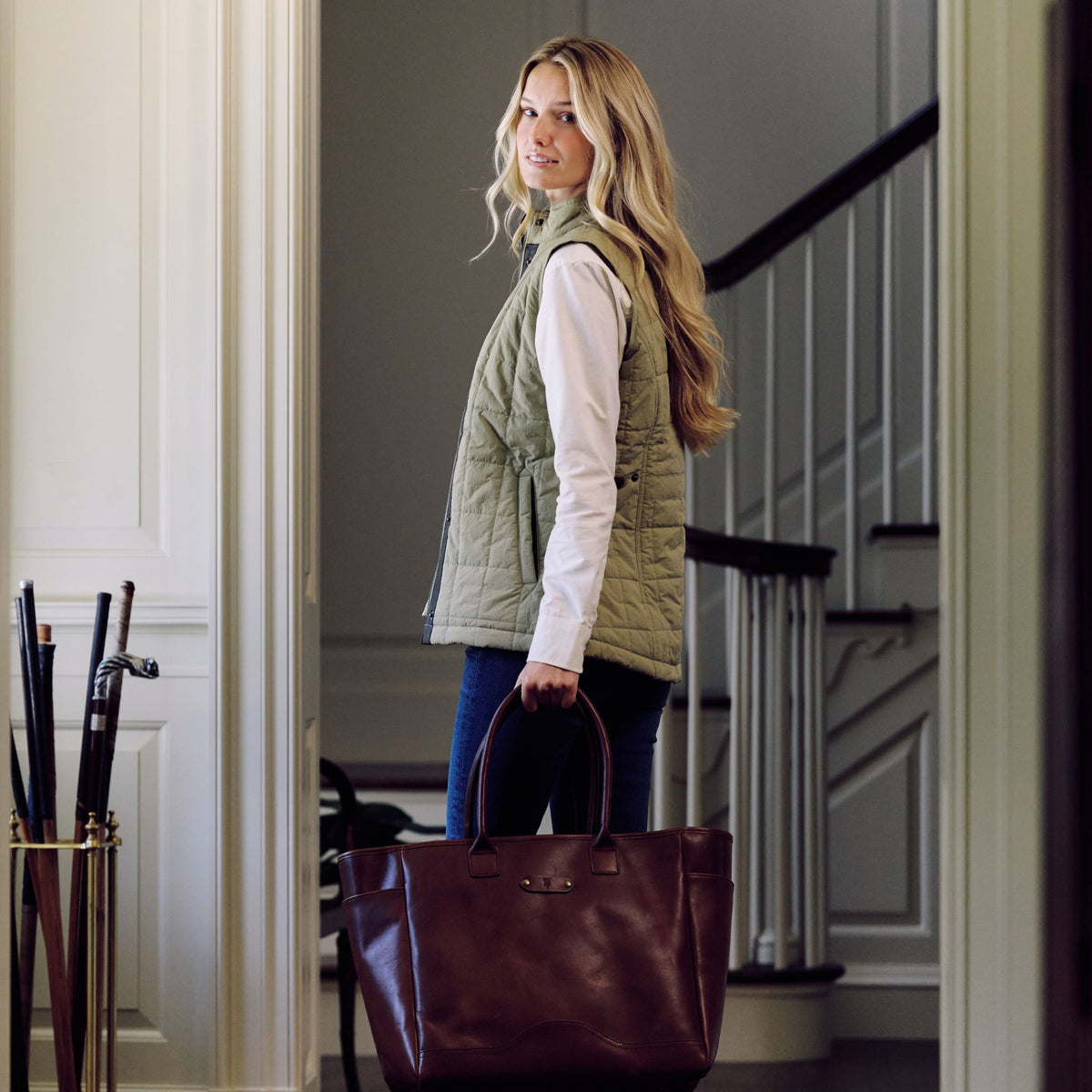 Woman stands indoors with stairs behind, wearing Tom Beckbe Women’s Tyler Quilted Vest, jeans, and holding a leather bag.