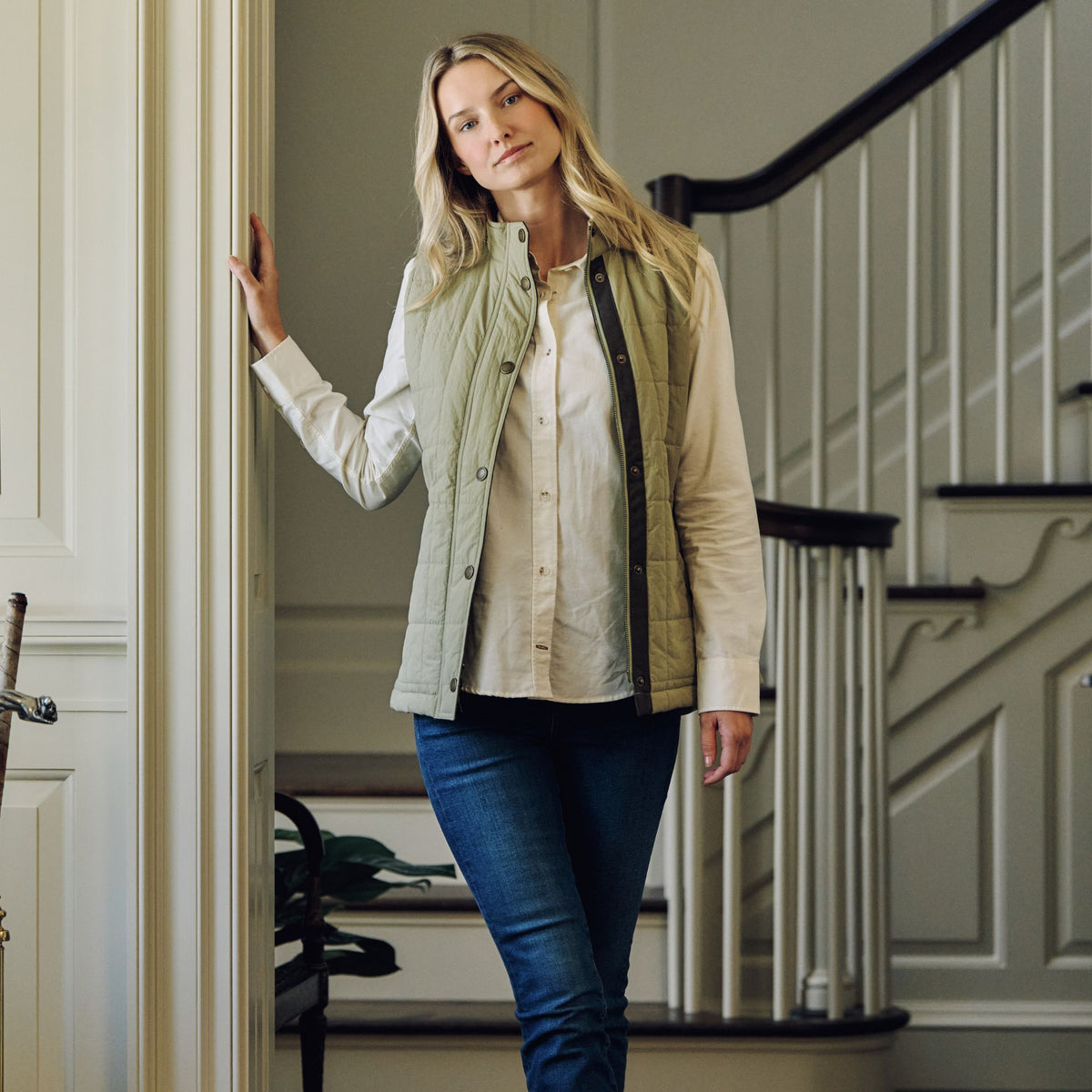 Woman in Tom Beckbe Women’s Tyler Quilted Vest and white shirt stands indoors by stairs, looking at the camera.