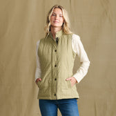 Woman in a Tom Beckbe Women’s Tyler Quilted Vest over a cream shirt and jeans, standing by a tan backdrop.