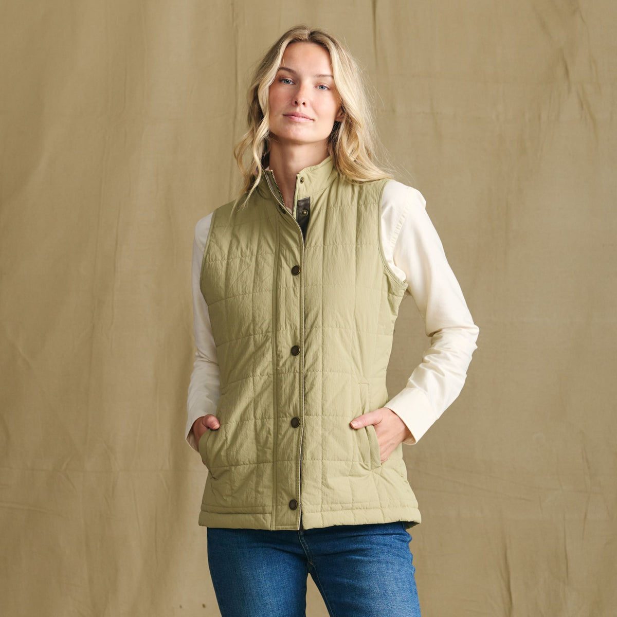Woman in a Tom Beckbe Women’s Tyler Quilted Vest over a cream shirt and jeans, standing by a tan backdrop.