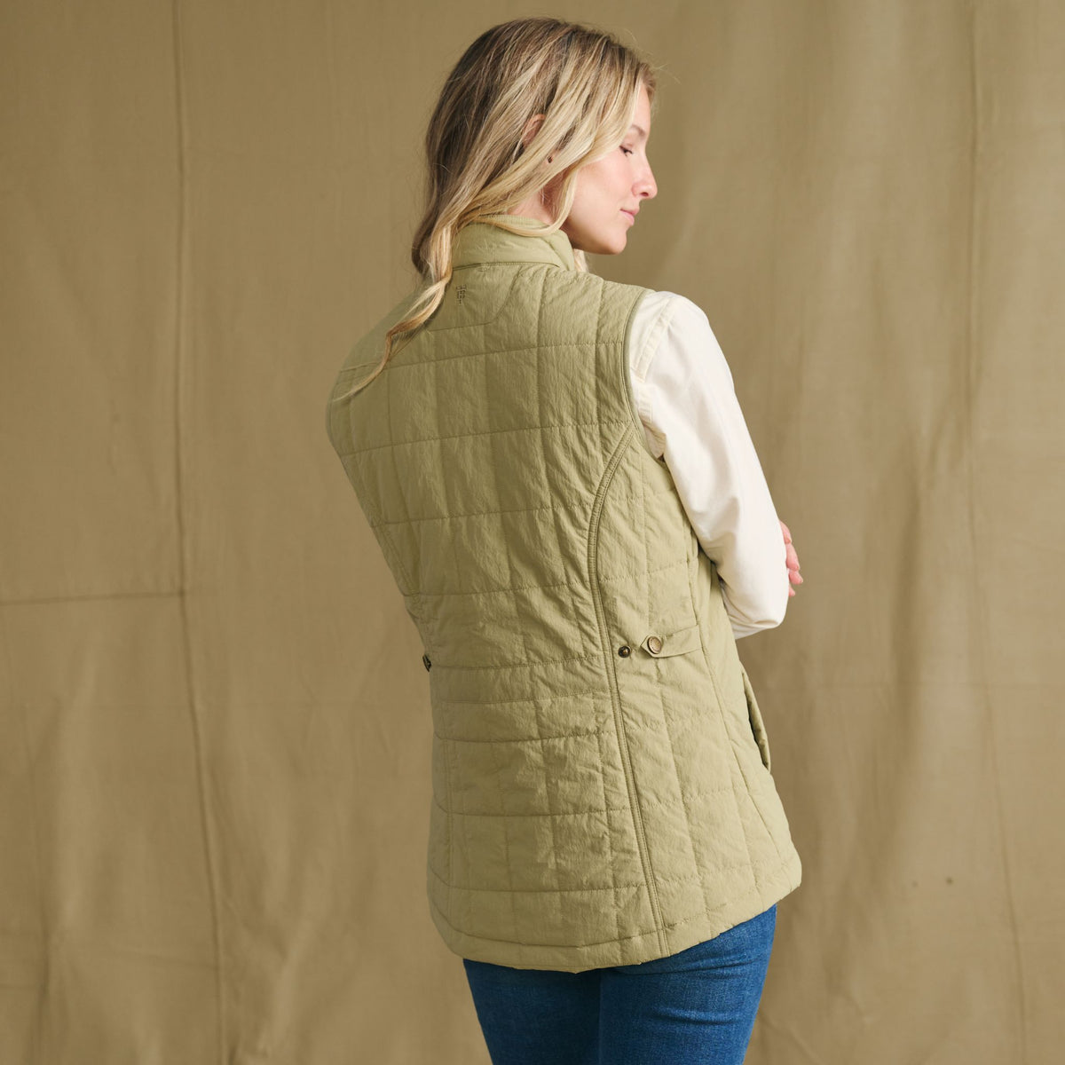 Woman wears Tom Beckbe Women’s Tyler Quilted Vest in green over white shirt, facing away against plain tan background.