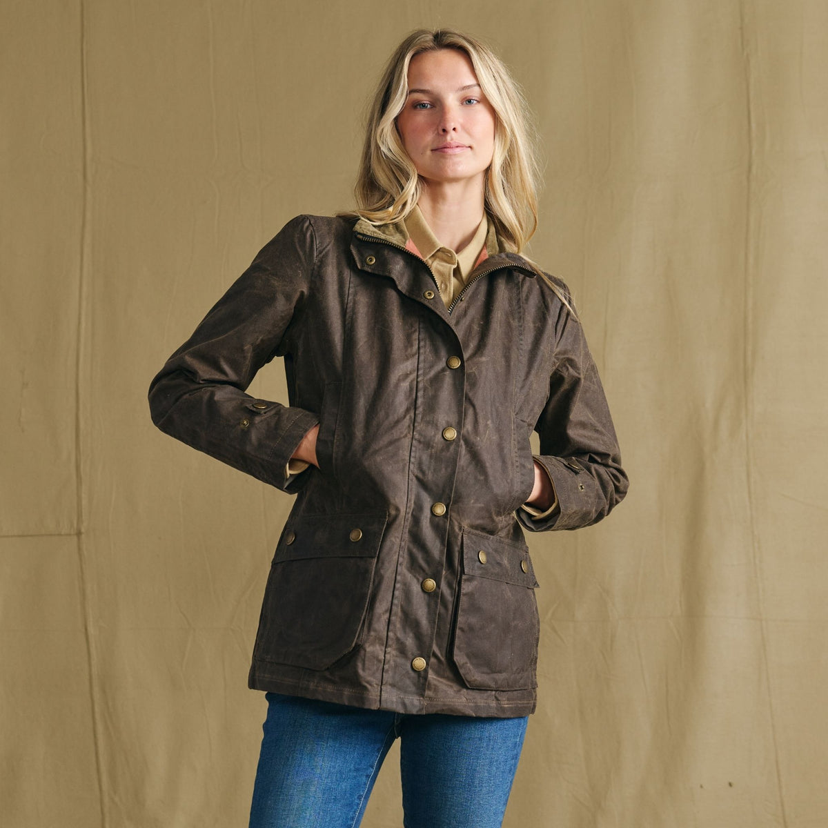 Woman in Tom Beckbe’s Women’s Blakeley Jacket and jeans stands by a tan fabric backdrop.
